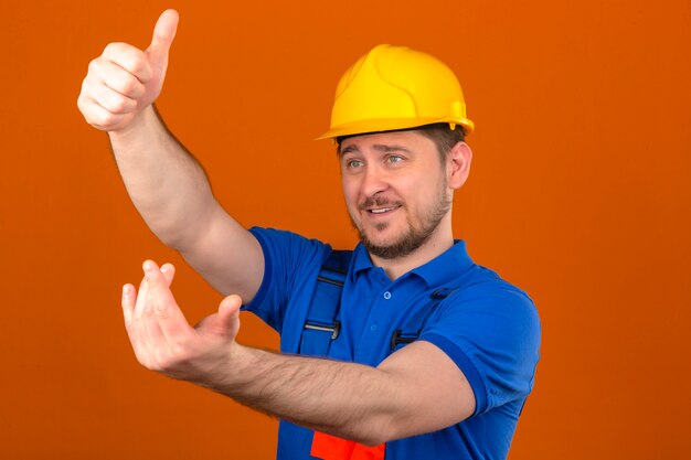 Budowniczy mężczyzna w mundurze konstrukcyjnym i hełmie ochronnym, zapraszający do zbliżenia się, wykonujący gest pozytywną i przyjazną ręką nad odizolowaną pomarańczową ścianą