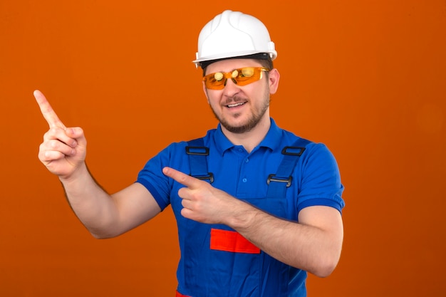 Budowniczy mężczyzna w okularach mundurowych i hełmie ochronnym, uśmiechnięty wesoło i wskazujący rękami i palcem w bok, stojąc nad izolowaną pomarańczową ścianą