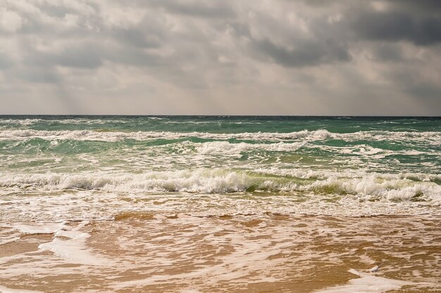 Burza na piaszczystej plaży wybrzeża Morza Czarnego, szmaragdowo czysta woda, ciepły dzień. Promienie słoneczne przedzierają się przez chmury na niebie.