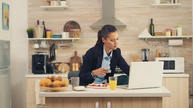 Businesswoman dokonywania płatności online za pomocą karty kredytowej na laptopie podczas śniadania. Zakupy online za towary i ubrania, korzystanie z nowoczesnych technologii w życiu codziennym, dokonywanie płatności przez internet