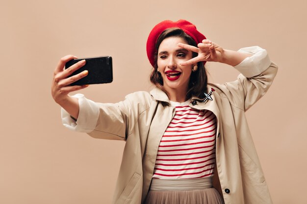 Całkiem cudowna kobieta o brązowych włosach w czerwonym kapeluszu mrugająca, pokazująca znak pokoju i biorąca selfie na beżowym na białym tle.