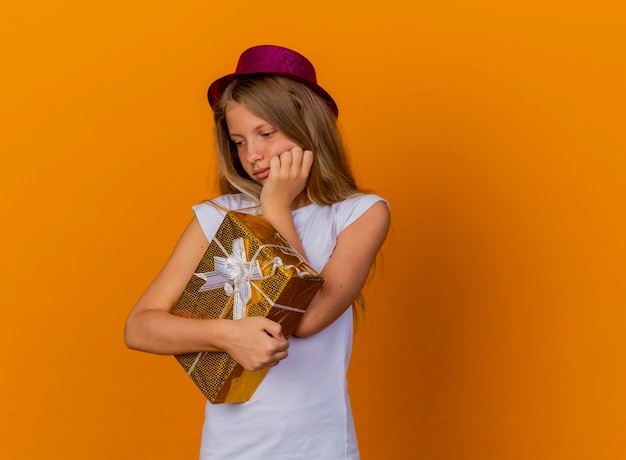 Bezpłatne zdjęcie całkiem mała dziewczynka w świątecznym kapeluszu trzyma pudełko, patrząc na bok z zamyślonym wyrazem myśli, koncepcja przyjęcie urodzinowe stoi na pomarańczowym tle