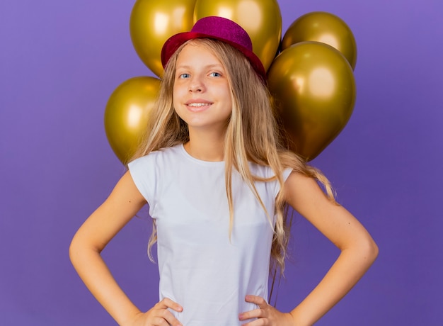 Całkiem mała dziewczynka w świątecznym kapeluszu z bukietem balonów patrząc na kamery, uśmiechając się radośnie, urodziny koncepcja stojąca na fioletowym tle