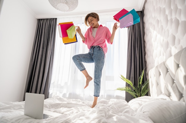 Całkiem szczęśliwa kobieta bawi się skacząc na łóżku w domu z kolorowymi torbami na zakupy