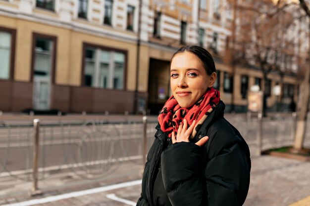 Całkiem urocza dziewczyna o ciemnych włosach, ubrana w czarną kurtkę i kolorowy szalik pozuje do kamery ze szczęśliwym uśmiechem podczas spaceru po mieście w promieniach słońca Ciepły słoneczny zimowy dzień w mieście