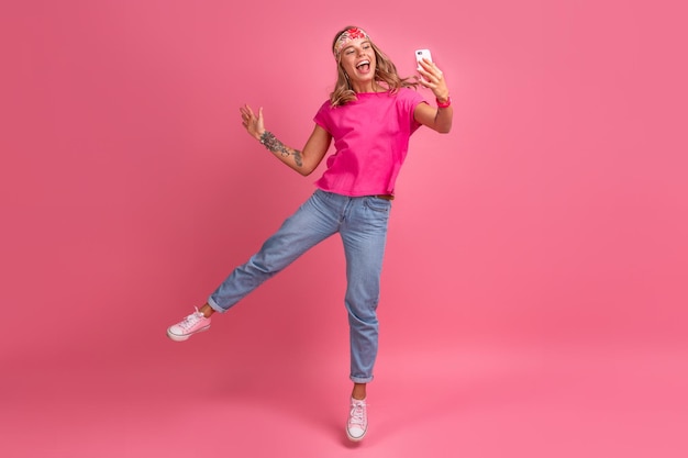 Bezpłatne zdjęcie całkiem urocza uśmiechnięta kobieta w różowej koszuli akcesoria w stylu boho hippie uśmiechnięta emocjonalna zabawa pozuje na różowym tle na białym tle pozytywny nastrój skaczący trzymając telefon