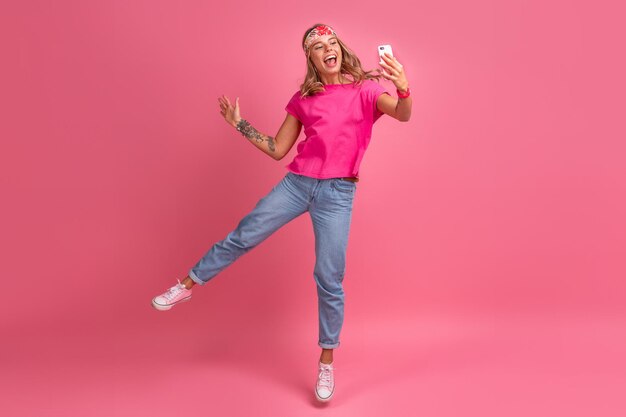 Całkiem urocza uśmiechnięta kobieta w różowej koszuli akcesoria w stylu boho hippie uśmiechnięta emocjonalna zabawa pozuje na różowym tle na białym tle pozytywny nastrój skaczący trzymając telefon