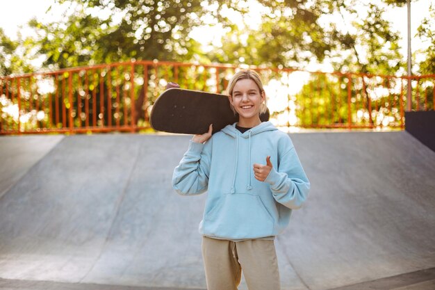 Całkiem uśmiechnięta dziewczyna w bluzie z kapturem, trzymająca deskorolkę na ramieniu, szczęśliwie pokazująca gest kciuka w skateparku