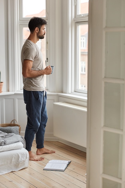 Całościowe ujęcie zamyślonego nieogolonego mężczyzny w swobodnym t-shircie i spodniach, stoi przy oknie i pije kawę