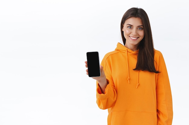 Charyzmatyczna uśmiechnięta młoda kaukaska dziewczyna trzymająca telefon komórkowy i pokazująca aplikację smartfona na wyświetlaczu