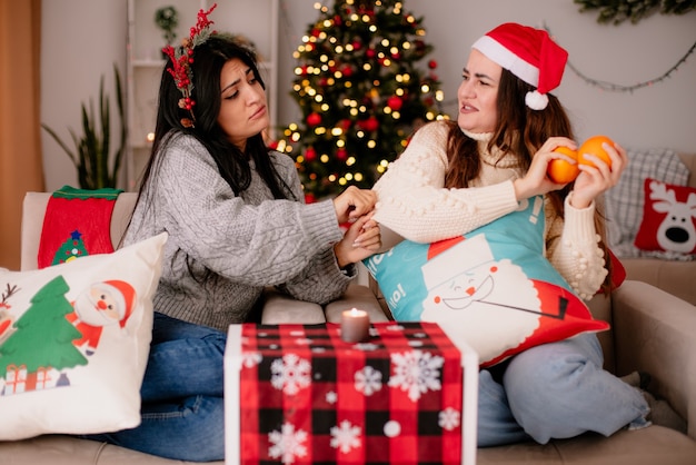 chciwa ładna młoda dziewczyna w czapce Mikołaja trzyma pomarańcze i patrzy na swojego przyjaciela z wieńcem ostrokrzewu siedzącego na fotelu Boże Narodzenie w domu