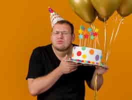 Bezpłatne zdjęcie chciwy dorosły słowiański mężczyzna w okularach optycznych w czapce urodzinowej z wystającym językiem trzyma balony z helem i tort urodzinowy