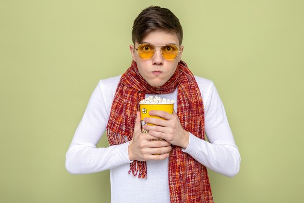 Chciwy młody przystojny facet ubrany w szalik w okularach, trzymający wiadro popcornu na oliwkowozielonej ścianie