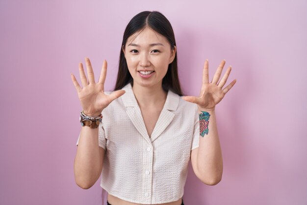 Chińska młoda kobieta stojąca na różowym tle, pokazująca i wskazująca palcami numer dziesięć, uśmiechając się pewnie i szczęśliwie.