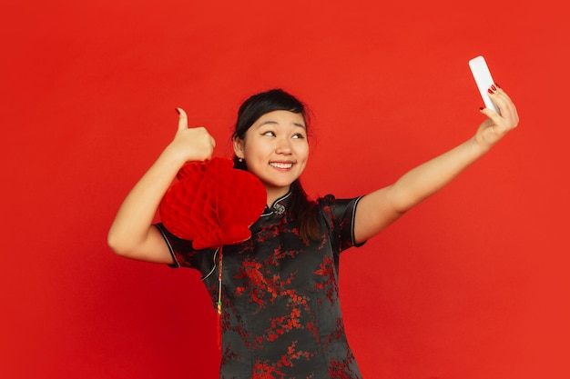 Chiński Nowy Rok. Portret młodej dziewczyny Azji na białym tle na czerwonym tle. Modelka w tradycyjnych strojach wygląda na szczęśliwą i robi selfie z dekoracją. Uroczystość, święto, emocje.