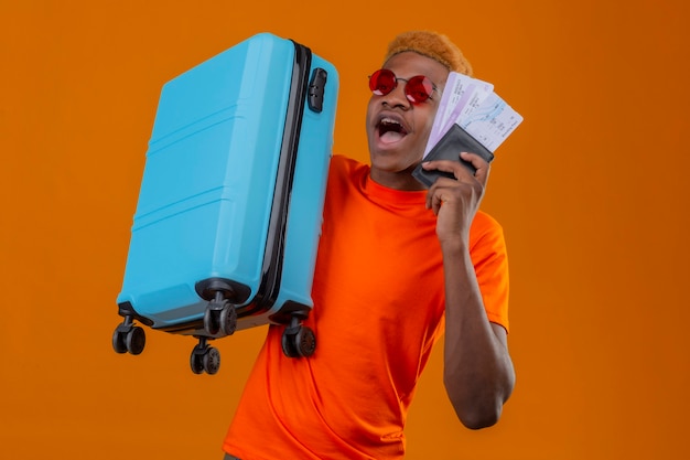 Chłopiec młody podróżnik ubrany w pomarańczową koszulkę, trzymając walizkę i bilety lotnicze, patrząc zdziwiony i zaskoczony stojąc nad pomarańczową ścianą