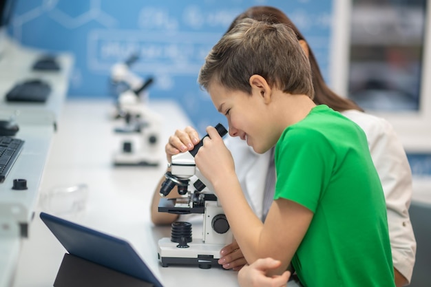Chłopiec patrzący przez mikroskop w pobliżu siedzącego nauczyciela
