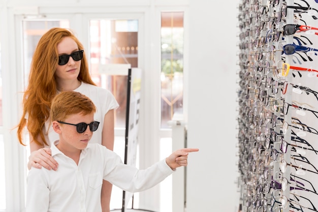 Chłopiec stoi z siostrą w sklepie optyka i wskazując na stojak na okulary