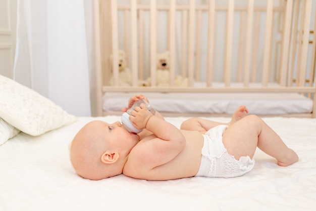 Chłopiec w wieku 8 miesięcy leżący w pieluchach na białym łóżku z butelką mleka w domu nogi do góry, koncepcja jedzenia dla niemowląt, woda do picia dla niemowląt z butelki