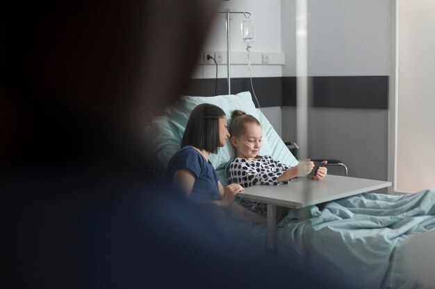 Chora dziewczynka odpoczywa na oddziale pediatrii szpitalnej podczas grania w gry na nowoczesnym telefonie komórkowym. Matka siedzi obok hospitalizowanej córki w trakcie leczenia, oglądając bajki na smartfonie.