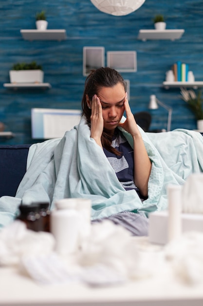 Chora kobieta z bólem głowy, siedząca na kanapie w domu, zażywająca tabletki i lecząca się z powodu przeziębienia grypy. Osoba z chorobami i problemami zdrowotnymi mająca migrenę i cierpiąca na objawy wirusa