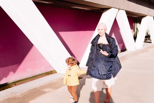 Chora na raka kobieta podająca sobie ręce z trzyletnim synkiem i nosząca dziecko w chuście idąc ulicą z różową ścianą w tle