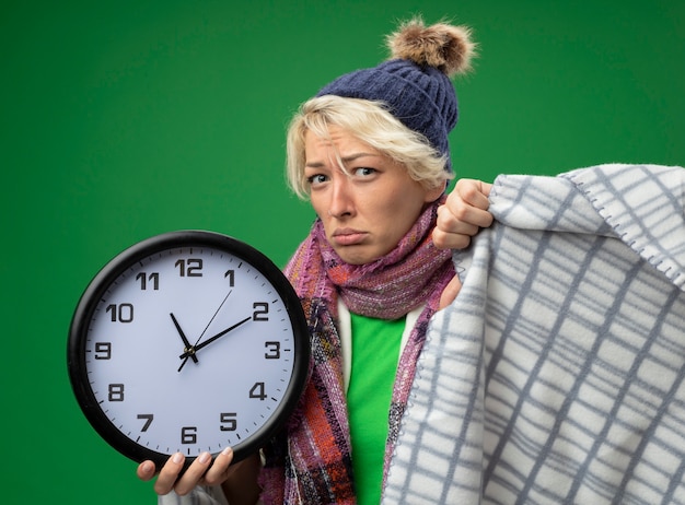 Bezpłatne zdjęcie chora niezdrowa kobieta z krótkimi włosami w ciepłym szaliku i czapce źle się czuje owinięta kocem trzymając zegar ścienny patrzy na kamerę z poważnym zielonym tłem