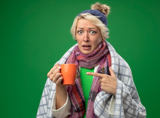 Bezpłatne zdjęcie chora, niezdrowa kobieta z krótkimi włosami w ciepłym szaliku i czapce źle się czuje owinięta w blank, zdezorientowana trzymająca filiżankę herbaty wskazująca z figneratem wskazującym na zielonym tle