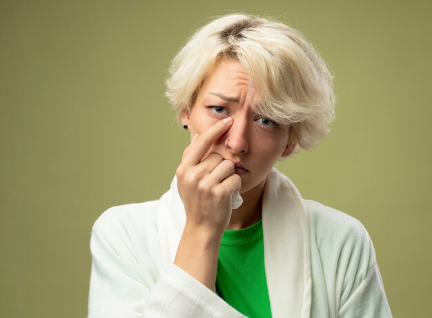 Chora niezdrowa kobieta z krótkimi włosami źle się czuje, wskazując palcem na nos, będąc nieszczęśliwą stojąc nad jasną ścianą
