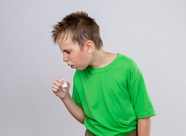 Chory chłopiec w zielonej koszulce czuje się niedobrze, kaszle i stoi nad białą ścianą