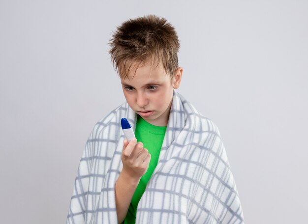 Chory chłopiec w zielonej koszulce zawiniętej w koc czuje się źle, patrząc na termometr zmartwiony stojąc nad białą ścianą