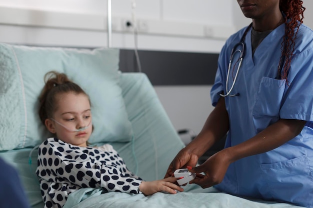 Chory dzieciak siedzi na szpitalnym oddziale pediatrycznym, podczas gdy personel medyczny monitoruje stan zdrowia za pomocą pulsoksymetru. Pielęgniarka mierząca poziom tlenu hospitalizowanej chorej małej dziewczynki odpoczywającej na łóżku pacjenta