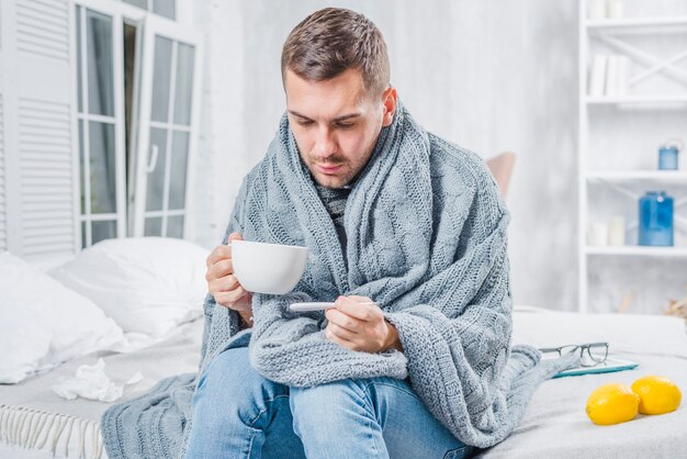 Chory mężczyzna obsiadanie na łóżku trzyma filiżankę kawy sprawdza gorączkę w termometrze