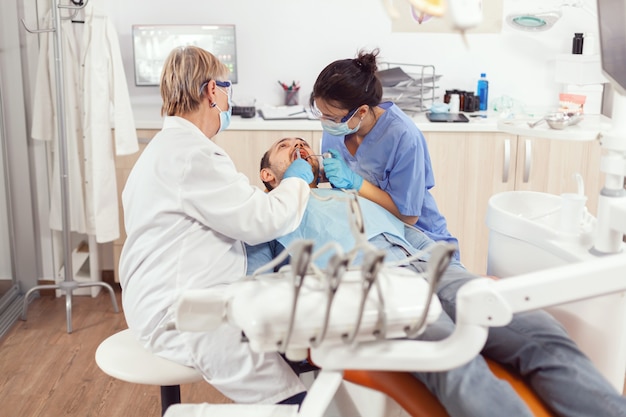 Chory pacjent siedzi na fotelu stomatologicznym z otwartymi ustami do wglądu, w gabinecie stomatologicznym, lekarz przygotowujący się do chirurgii jamy ustnej. dentysta i pielęgniarka medyczna z maskami usuwa ząb