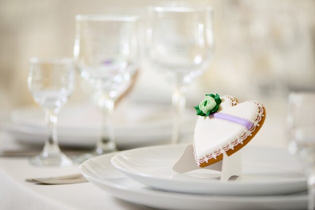 Ciastko w kształcie serca pokryte słodką glazurą, ozdobione zielonym kwiatkiem i drobnym wzorem, stoi na białych talerzach jako dekoracja świątecznego stołu weselnego przy kieliszkach
