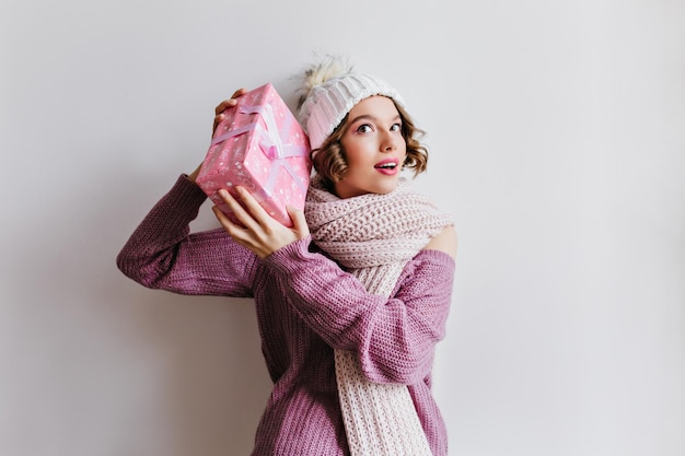 Ciekawa biała kobieta w długim szaliku z dzianiny, trzymająca prezent świąteczny w różowym pudełku. Beztroska dziewczyna w zimowym kapeluszu pozowanie na jasnym tle z prezentem na nowy rok.