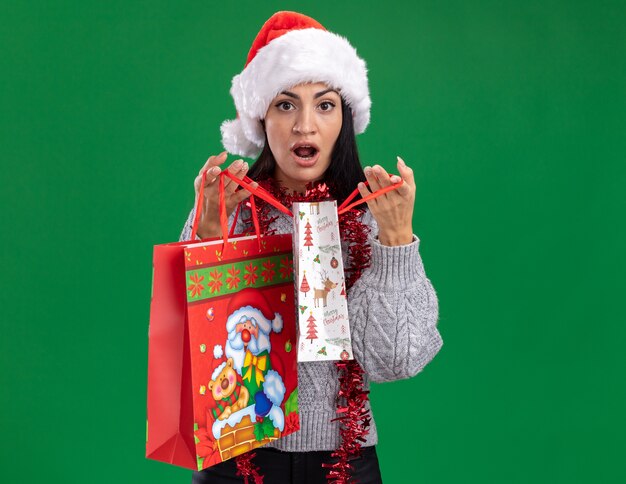 Ciekawa młoda kaukaska dziewczyna ubrana w świąteczny kapelusz i świecącą girlandę na szyi, trzymając torby na prezenty świąteczne, otwierając jedną na zielonej ścianie z miejscem na kopię
