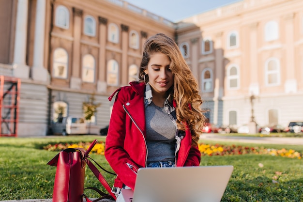 Ciekawa studentka w szarym swetrze siedzi na trawie z komputerem