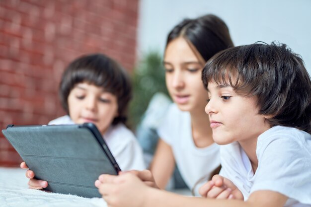 Ciekawy mały łaciński chłopiec patrząc na ekran komputera typu tablet. brat spędza czas z rodzeństwem, grając w gry, leżąc na łóżku w domu. szczęśliwe dzieci, koncepcja technologii. selektywne skupienie