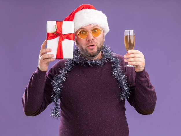 Ciekawy mężczyzna w średnim wieku w czapce świętego mikołaja i girlandzie świecidełka na szyi z okularami trzymającymi pakiet prezentów w pobliżu głowy i kieliszek szampana na fioletowej ścianie