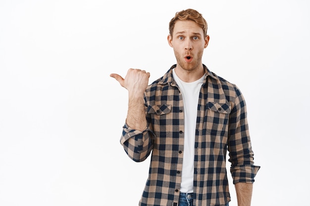 Ciekawy mężczyzna zadający pytanie o baner, wskazujący palcem na logo, demonstrujący reklamę i mówiący „wow”, będąc pod wrażeniem tekstu promocyjnego, stojący nad białą ścianą