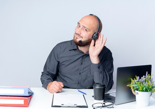 Ciekawy młody łysy mężczyzna call center sobie zestaw słuchawkowy siedzi przy biurku z narzędzi pracy robi nie słyszy, jak gest na białym tle