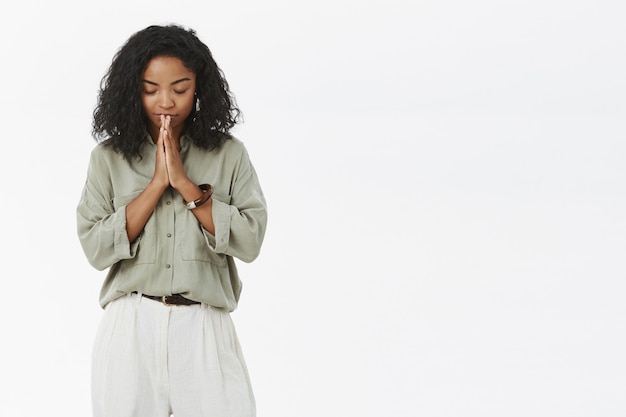 Ciemnoskóra kobieta pochylająca głowę zamykająca oczy stojąca spokojnie i zrelaksowana z rękami w modlitwie