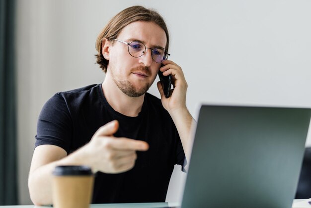 Cieszący się dobrym dniem pracy Pewny siebie młody człowiek pracuje na laptopie i rozmawia przez telefon komórkowy, siedząc w swoim miejscu pracy w biurze