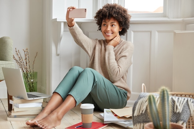 Cieszę się, że kobieta rasy mieszanej z fryzurą afro, pozuje do telefonu komórkowego do robienia selfie