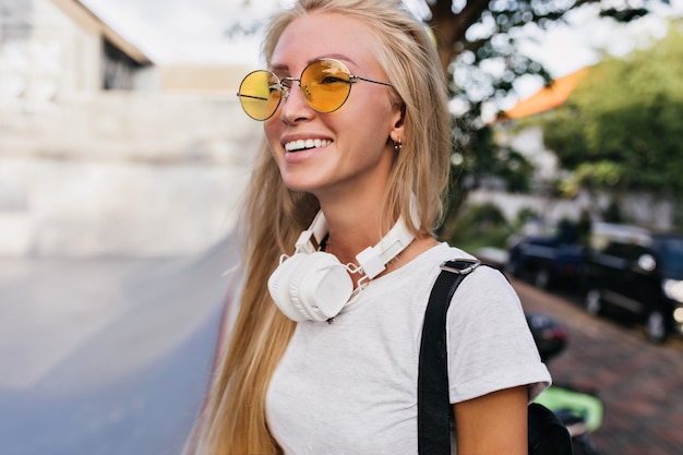Cieszę się, że młoda kobieta spaceruje po mieście w słuchawkach z wyrazem twarzy szczęśliwy. Ładna blond kobieta w żółte okulary, śmiejąc się podczas pozowania na rozmycie tła ulicy.