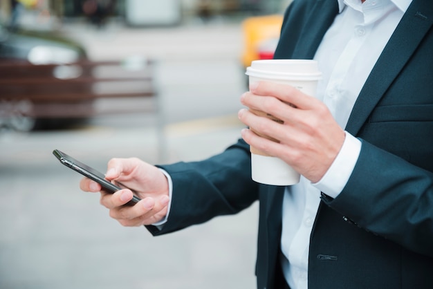 Close-up biznesmen ręki trzymającej filiżankę kawy na wynos za pomocą telefonu komórkowego