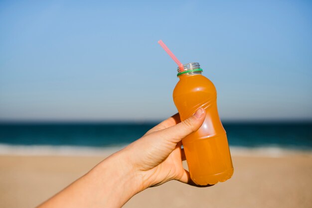 Close-up kobiecej ręki trzymającej plastikową butelkę soku pomarańczowego ze słomką do picia na plaży