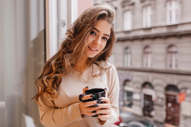 Close-up portret inspirowanej ciemnowłosej damy z dużymi oczami mrożącymi krew w żyłach na parapecie