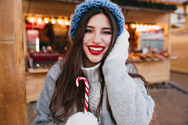 Close-up portret inspirowanej dziewczyny w ciepłych białych rękawiczkach pozujących z bożonarodzeniową laską.